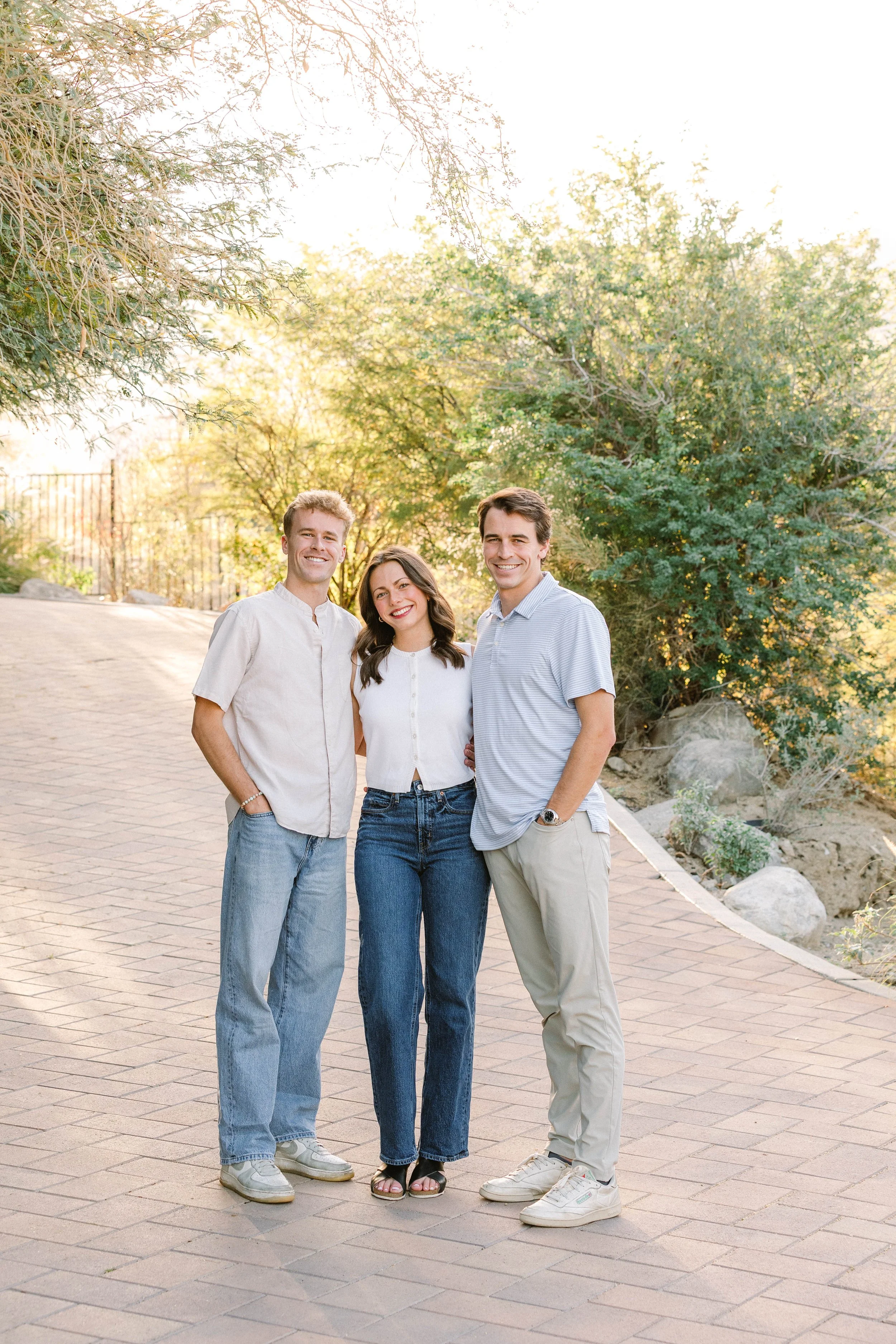 Three young adults standing on a brick path outdoors, smiling, with trees and rocks in the background during daylight.