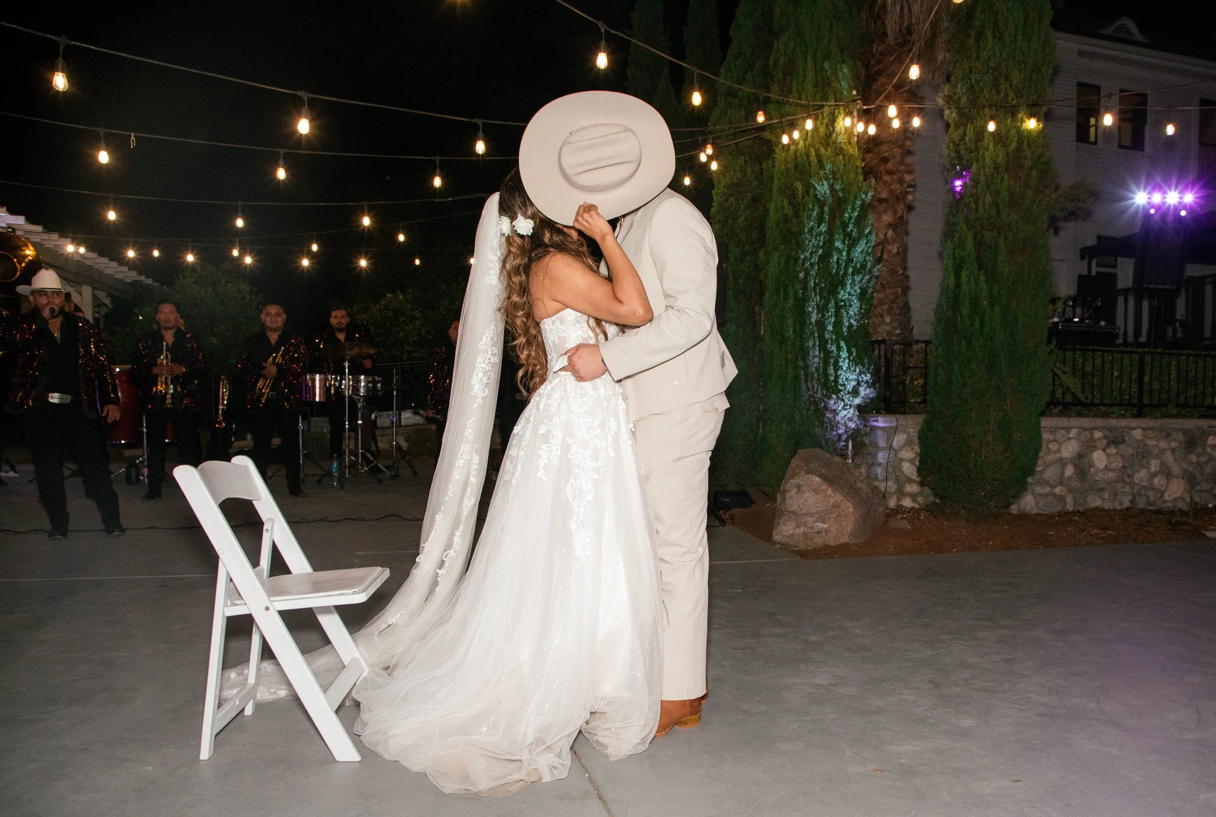 A bride and groom are dancing at their wedding reception, with the groom's face covered by a white hat, under string lights, with a band in the background and outdoor setting.