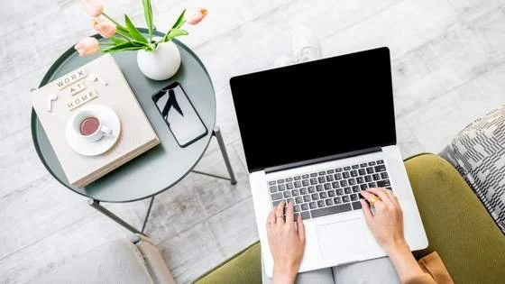 “Overhead view of a woman working remotely on a laptop with coffee, phone, and notebook on a side table with title "Work At Home."