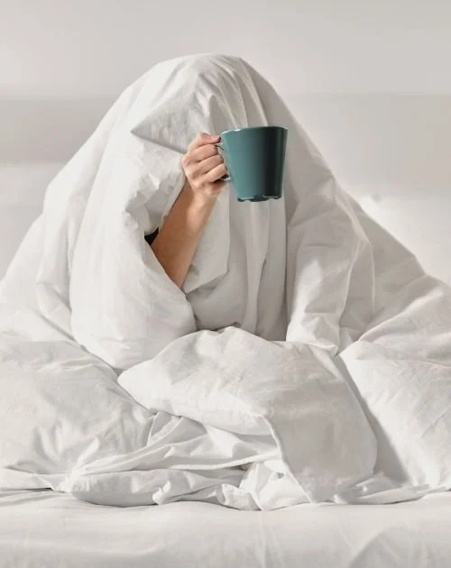 Exhausted woman sitting in bed under a white duvet holding a teal coffee mug