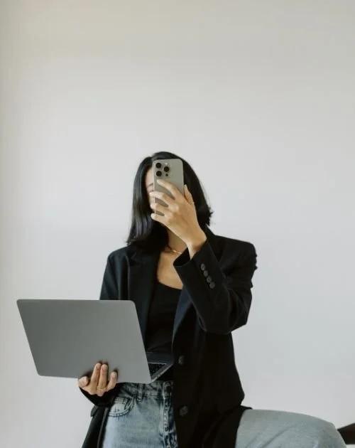 Professional woman in a black blazer holding an open laptop and taking a mirror selfie with her smartphone.