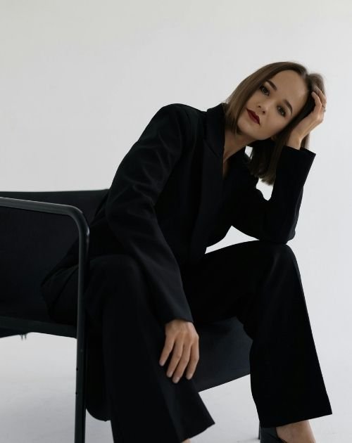 Woman in a black suit sitting on a chair, leaning forward with her head resting on her hand appearing stressed.