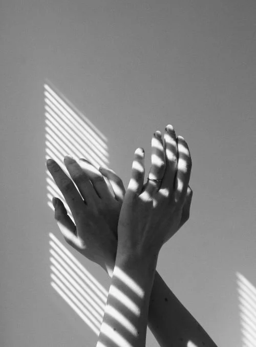 Black and white photo of two hands raised upward with light and shadow striped patterns across them against a light background.
