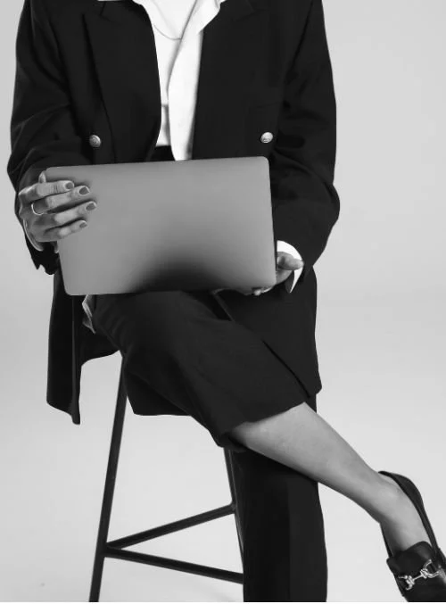 Woman in a black blazer sitting and holding a closed laptop against her body, photographed from the shoulders down.