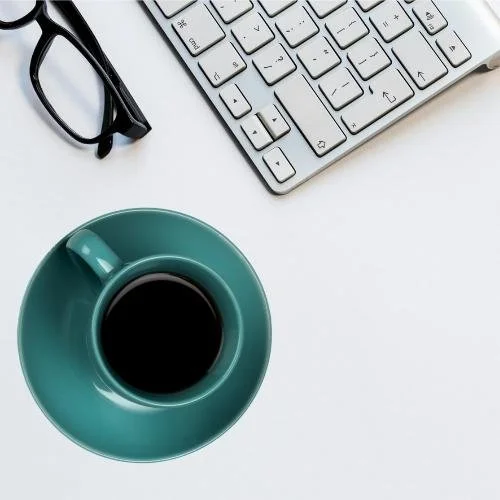 Teal coffee mug on white desk next to a pair of reading glasses and a mac keyboard.