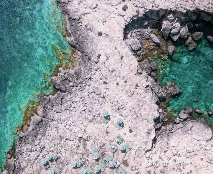 Aerial view of a rocky coastal area with calm turquoise water, a dock, and a group of small boats anchored near the shore.