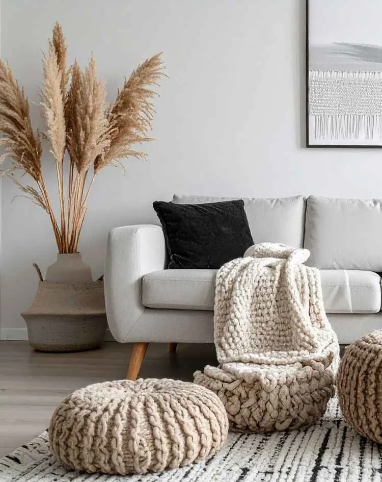 Living room with a light gray sofa, black throw pillow, chunky off-white knit blanket, large woven pouf, and a woven rug. A tall vase with dried pampas grass is on a side table next to the sofa. There is a framed abstract art piece on the wall.
