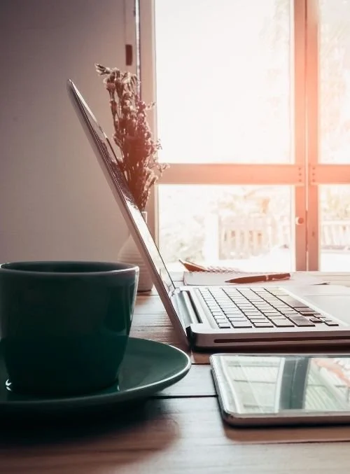 A laptop, a smartphone, a cup on a saucer, and a notebook on a wooden table near a window with sunlight streaming in.