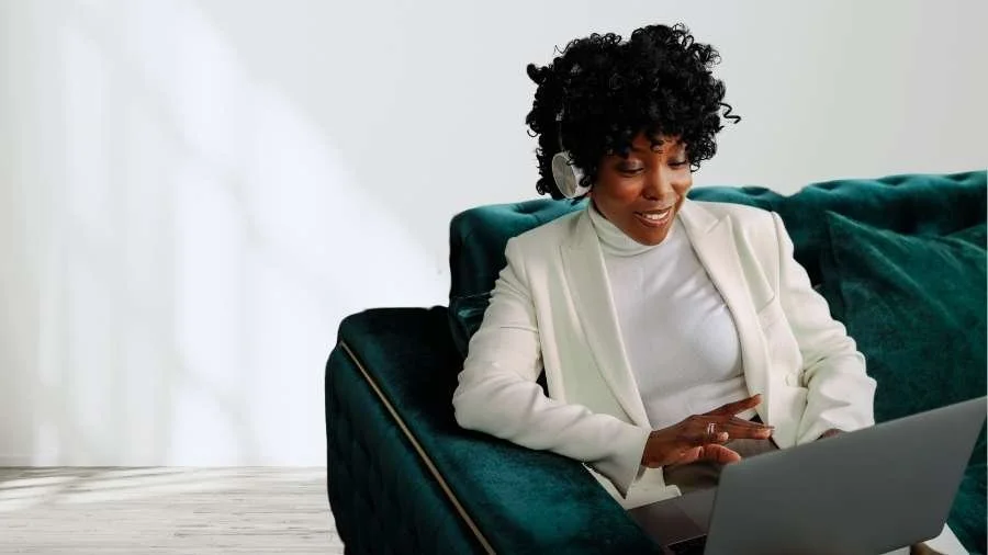 A woman with curly hair wearing headphones and a white blazer, sitting on a dark teal sofa, smiling at her laptop in a bright room.