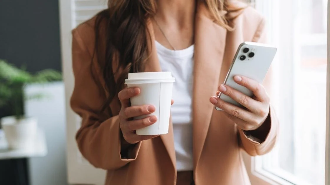 Boston professional in in tan blazer holding a coffee cup and checking her phone near a window