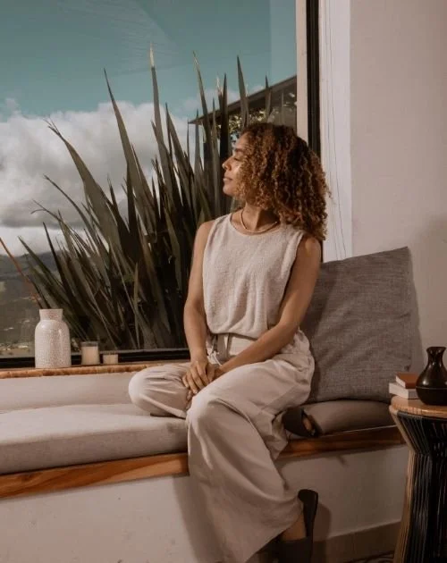 A woman with curly hair sits on a window seat, looking outside with a serene expression. She is wearing a sleeveless beige top and matching loose pants. Behind her are large plant leaves and a window showing a partly cloudy sky.