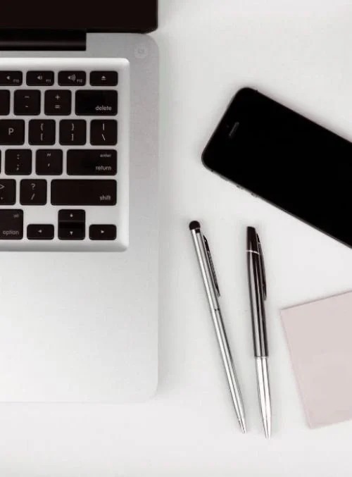 Close-up of a laptop keyboard, two pens, a smartphone, and a pink notepad on a white desk.