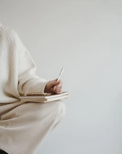 Person in a cream sweater writing in a notebook while seated, representing reflection and the therapy process