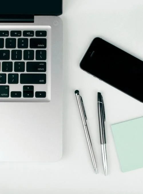 Minimal workspace with part of a laptop keyboard, black smartphone, two silver pens, and a light green notebook on a white desk
