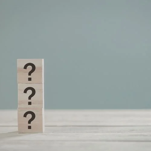 Three wooden blocks stacked vertically with black question marks on each, on a light-colored surface against a plain background.