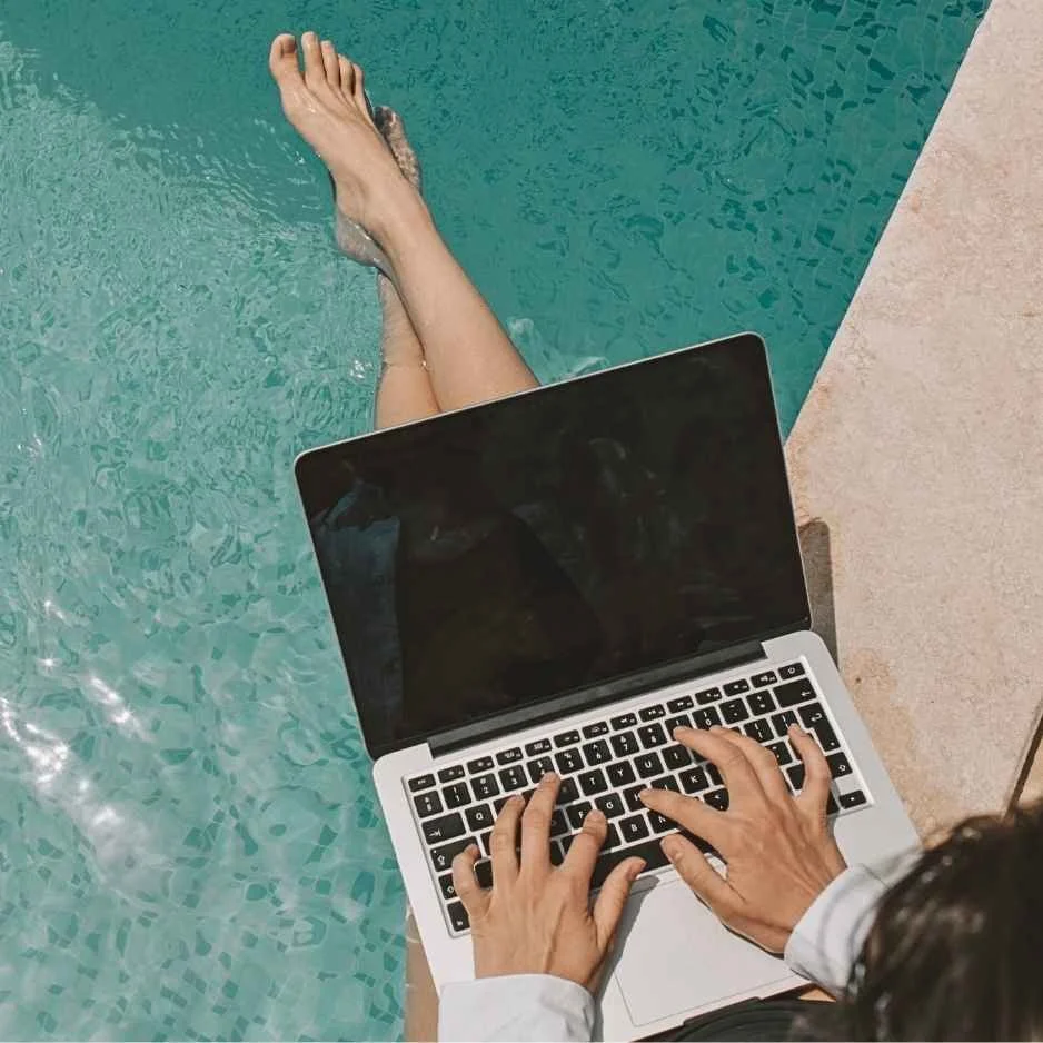 Female business owner working on a laptop by a swimming pool, illustrating work-life balance and mental health support for entrepreneurs.