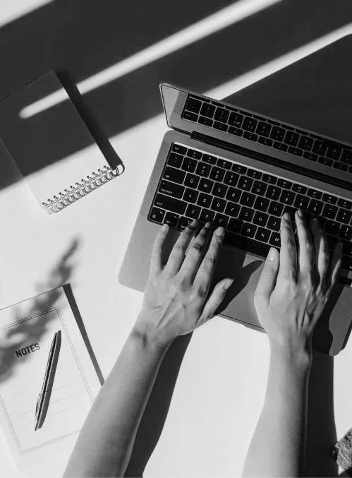 Overhead view of hands typing on a laptop next to an open spiral notebook on a white desk.