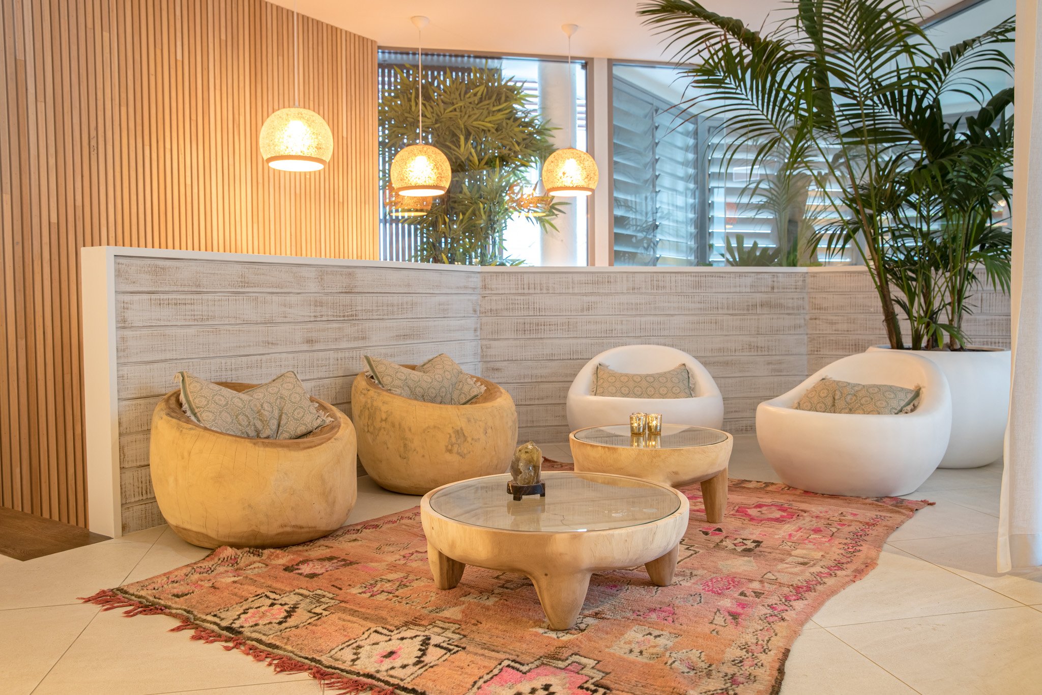 Modern lounge area with beige chairs, a round wooden coffee table, a pink patterned rug, large potted plants, and pendant lights hanging from the ceiling.