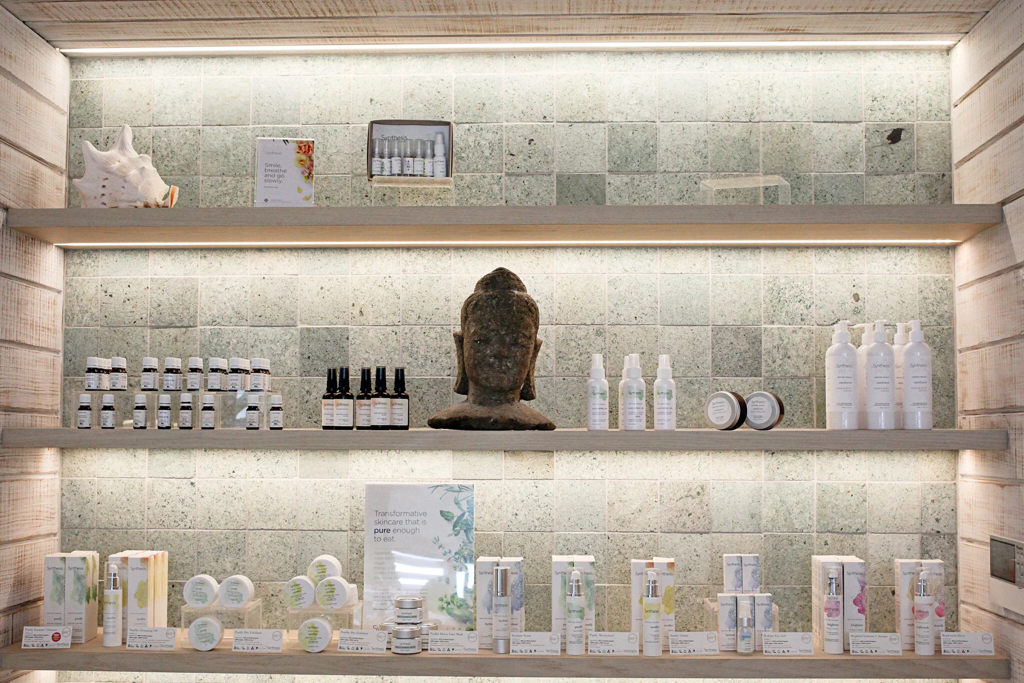 Spa or skincare product display with bottles, jars, and a Buddha head statue on wooden shelves against green tiled wall.