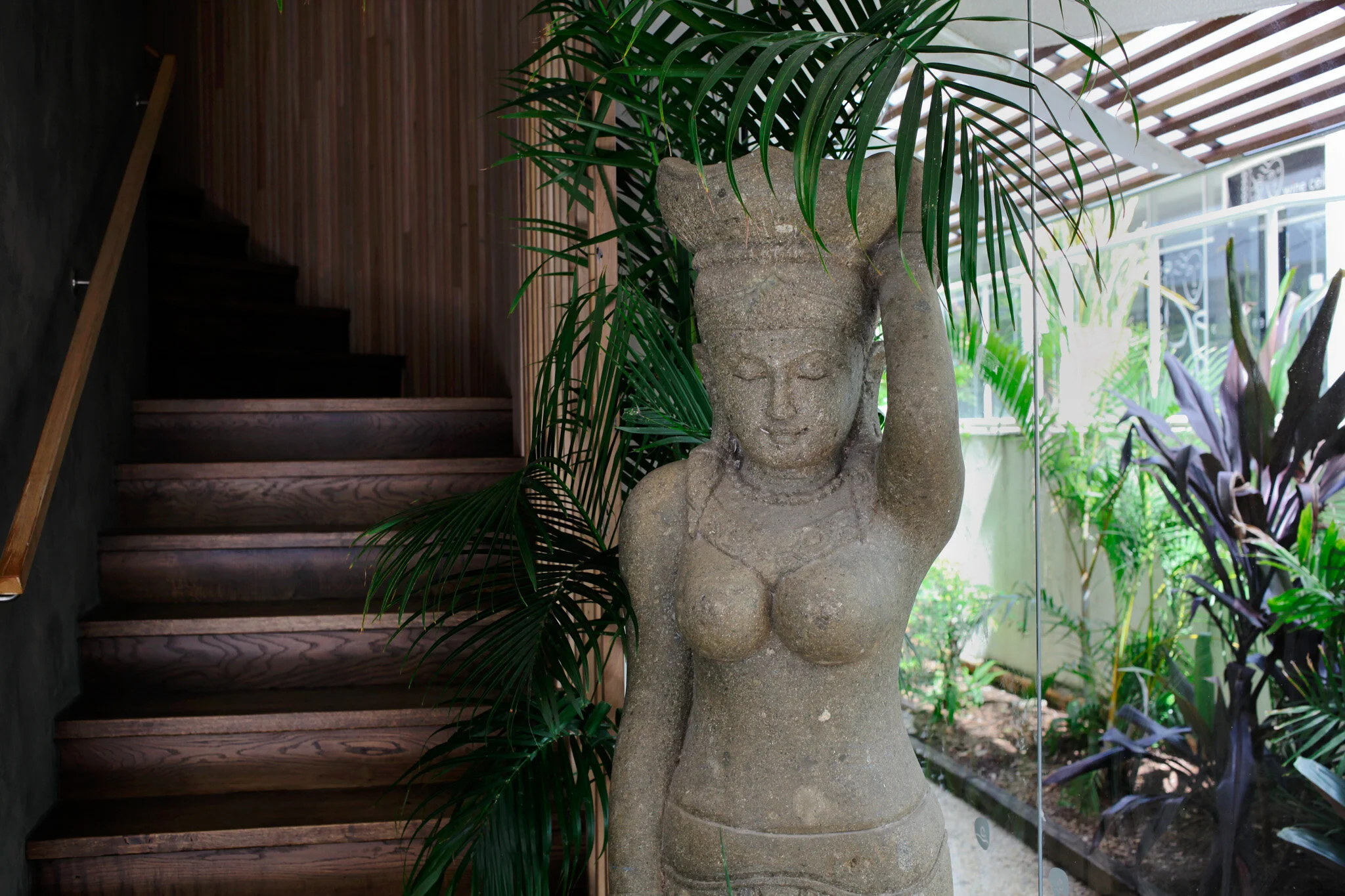 Stone sculpture of a woman holding a large bowl above her head, surrounded by houseplants.