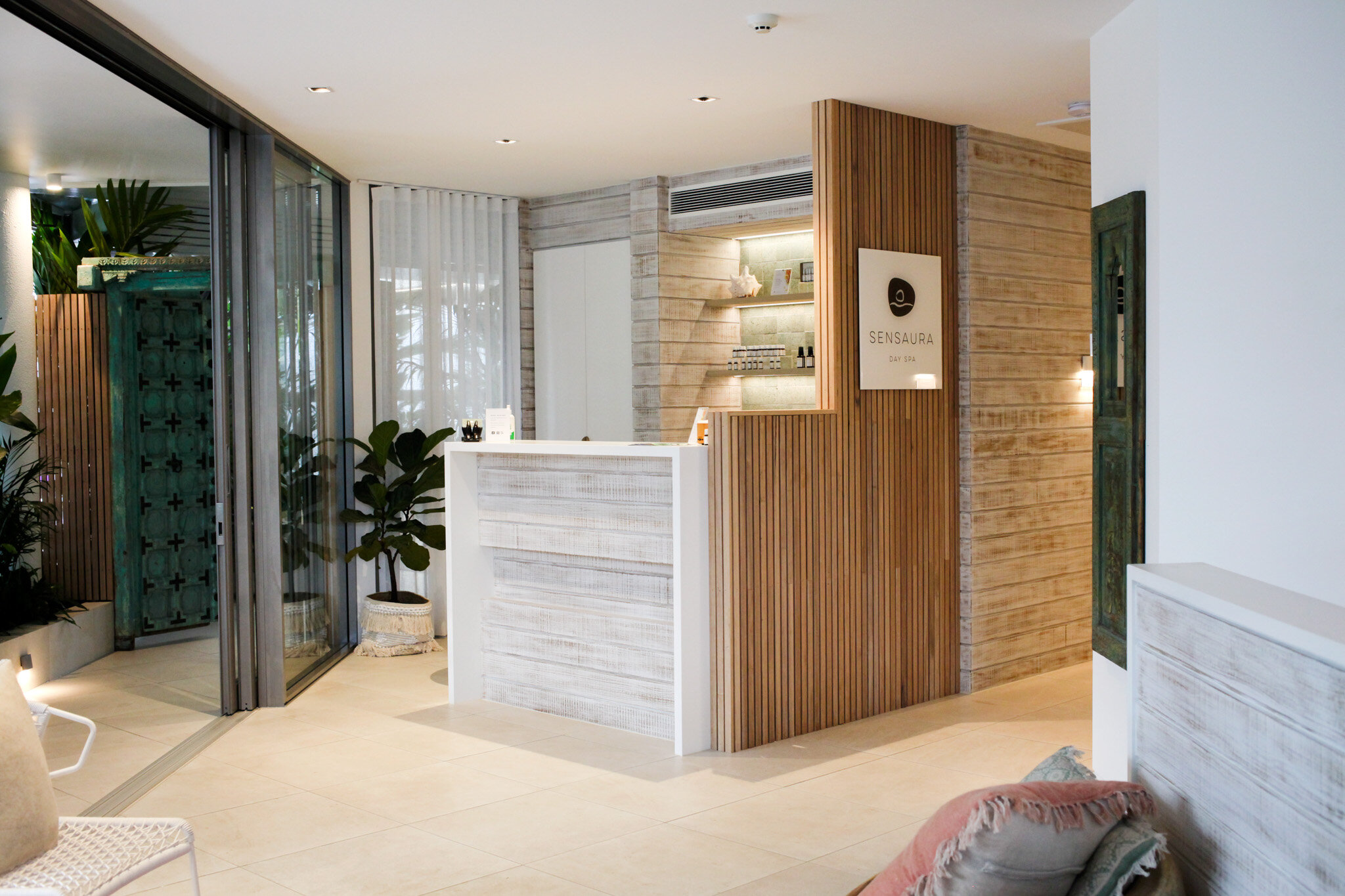 Spa reception area with a wooden desk, potted plant, and sign reading 'Sensaura Day Spa' on wooden wall.