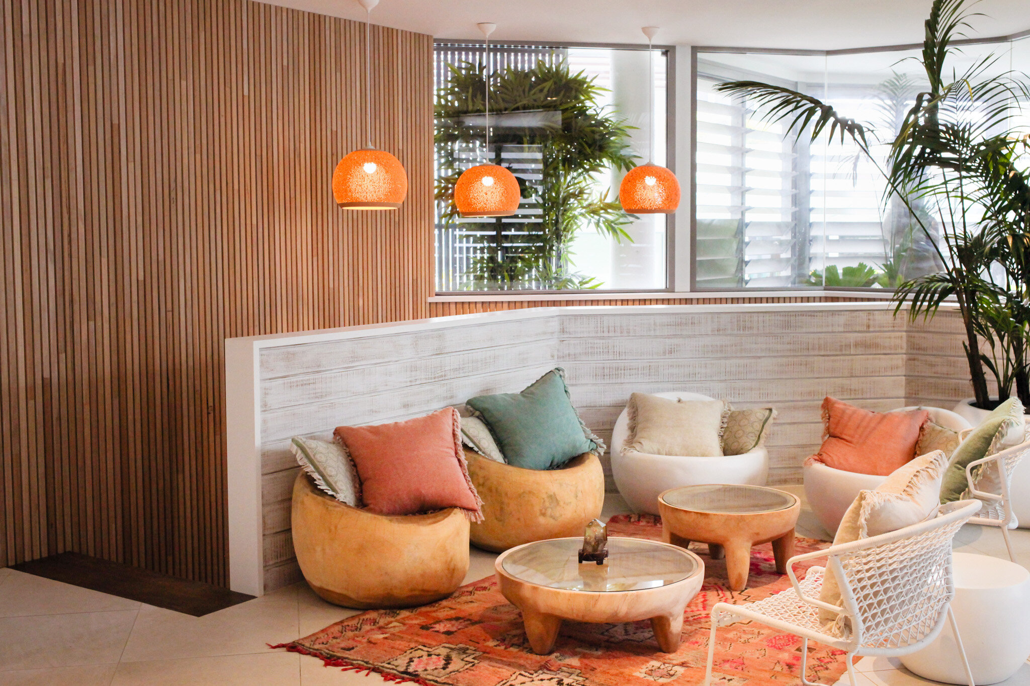 A cozy lounge area with cushioned chairs and round wooden tables, decorated with pillows, set on an orange patterned rug. There are three hanging orange pendant lights and large windows letting in natural light, with plants outside and a tall potted 