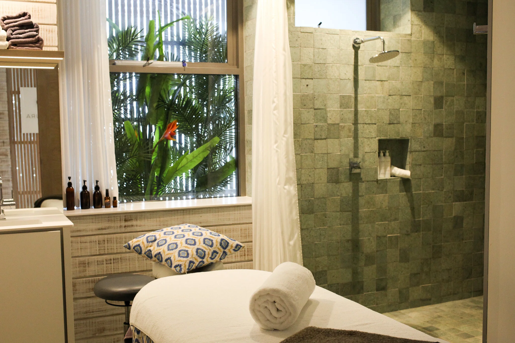 A spa or massage room with a bed covered in white towels, a pillow, and a brown towel. There is a small round stool and a window with curtains and green plants outside. The room has a glass shower area with a rainfall showerhead and shampoo bottles in a built-in niche.