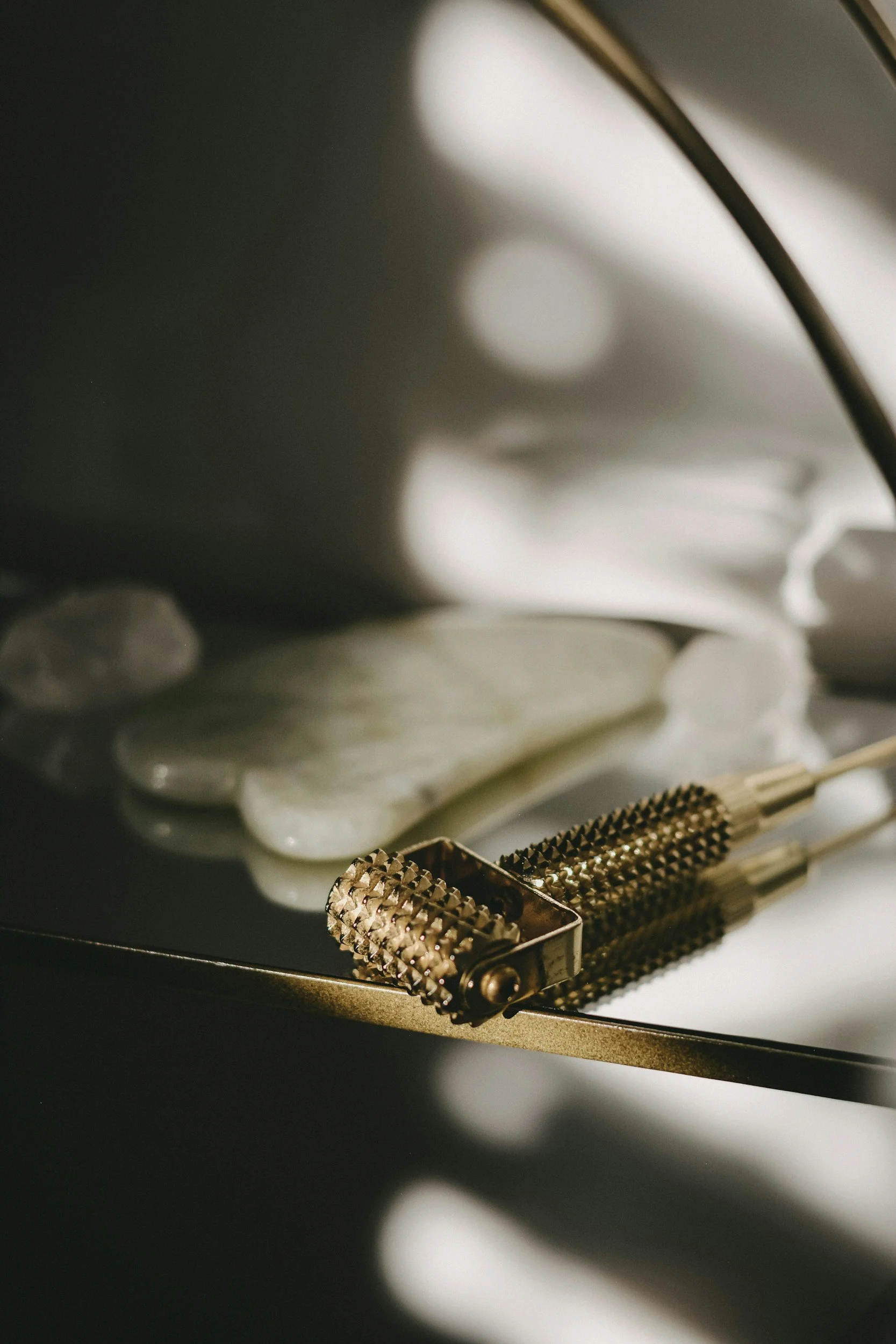 Close-up of metallic dermaroller tools with textured roller heads on a dark surface, with smooth white stones and blurred light circles in the background.