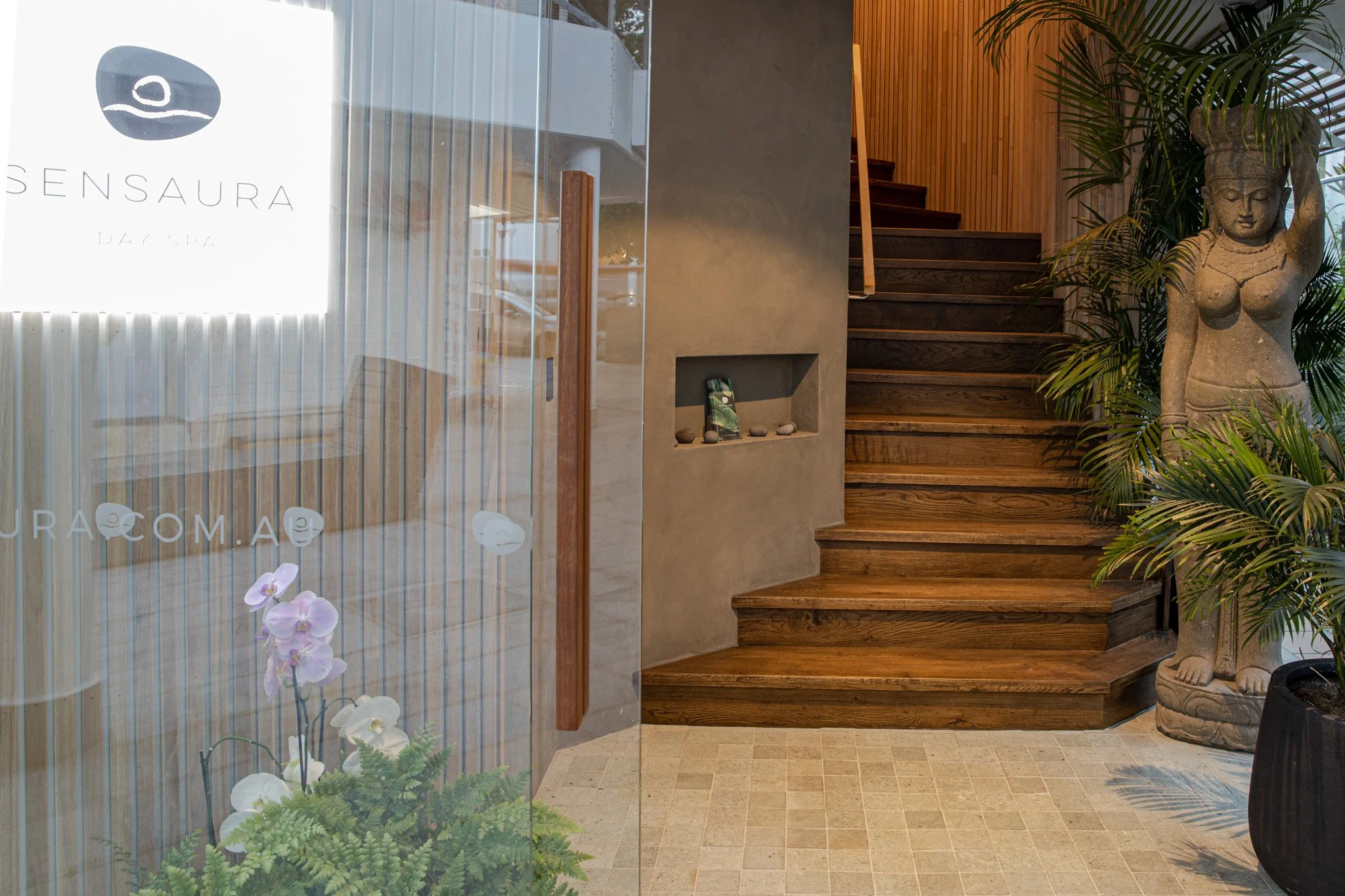 Interior of a spa with wooden stairs, a stone statue of a woman, lush green plants, and a glass wall with a sign that reads 'Sensuara Day Spa'.