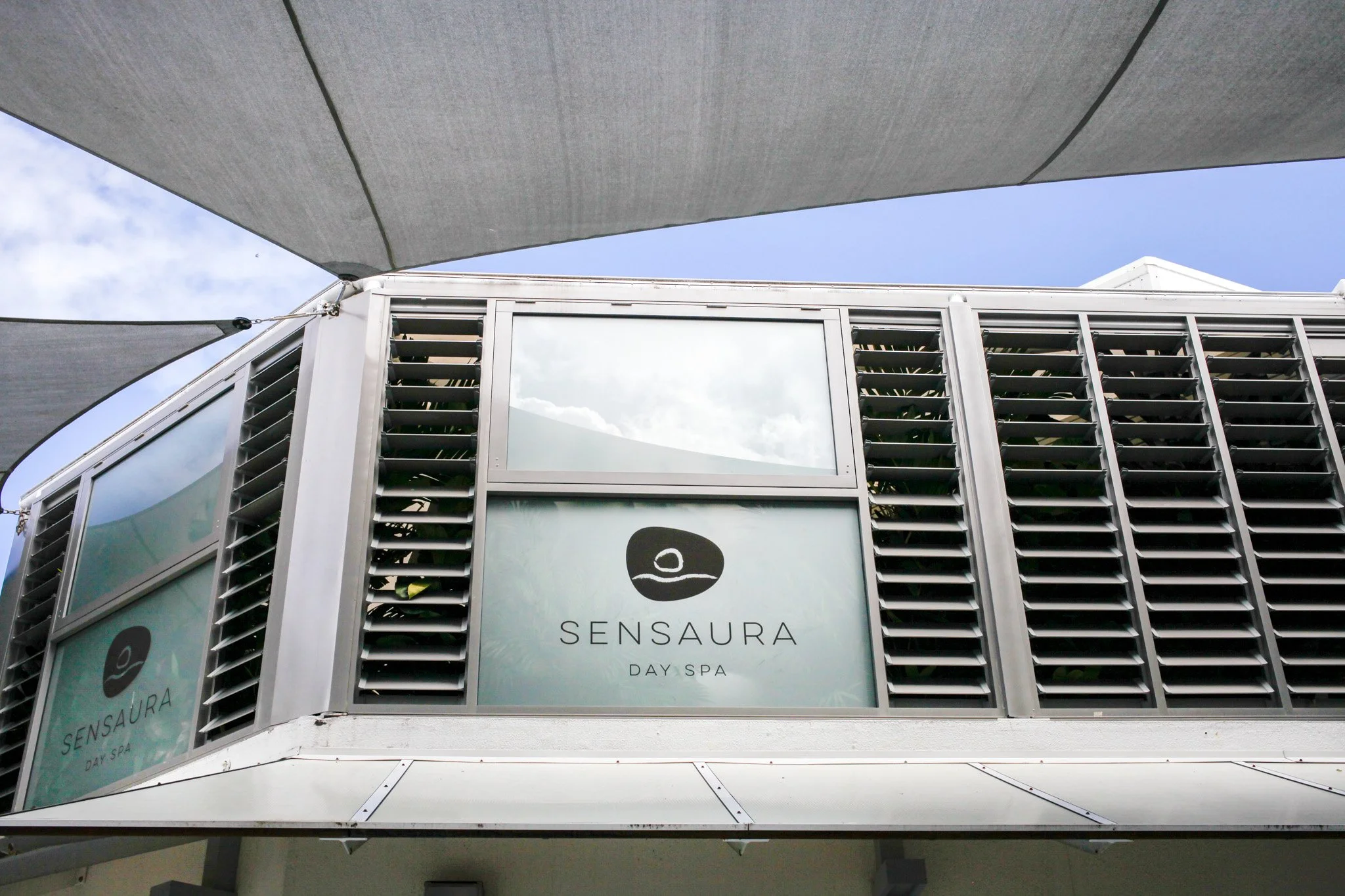 Exterior of a building with a sign that reads 'Sensaura Day Spa' and includes a circular logo, under a large shade cloth against a blue sky.