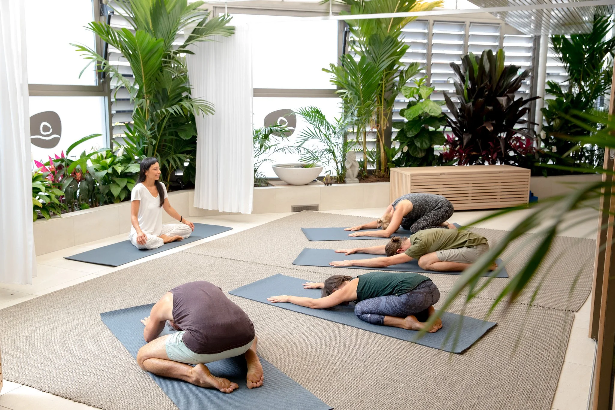 A group of people participating in a yoga class, kneeling on yoga mats with arms extended forward and heads down, following an instructor seated cross-legged on a separate mat in a room with large windows, abundant greenery, and decorative items.