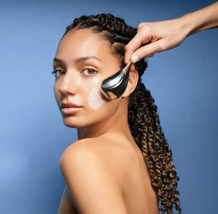 A woman with curly hair has cream applied to her cheek with a metal applicator as part of a skincare routine, against a blue background.
