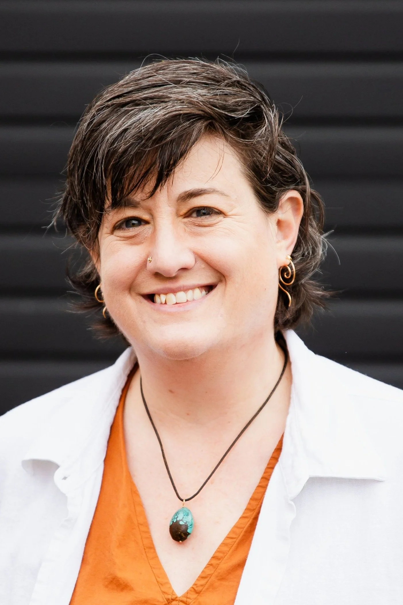 Close-up of a smiling person with short brown hair, hoop earrings, and a turquoise pendant necklace, wearing a white shirt over an orange top against a black background.
