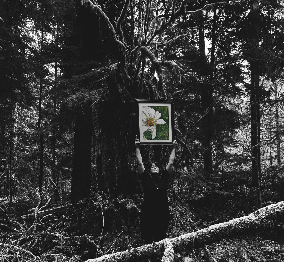 A person standing in a forest holding a framed picture of a white flower with a yellow center against a green background above their head.