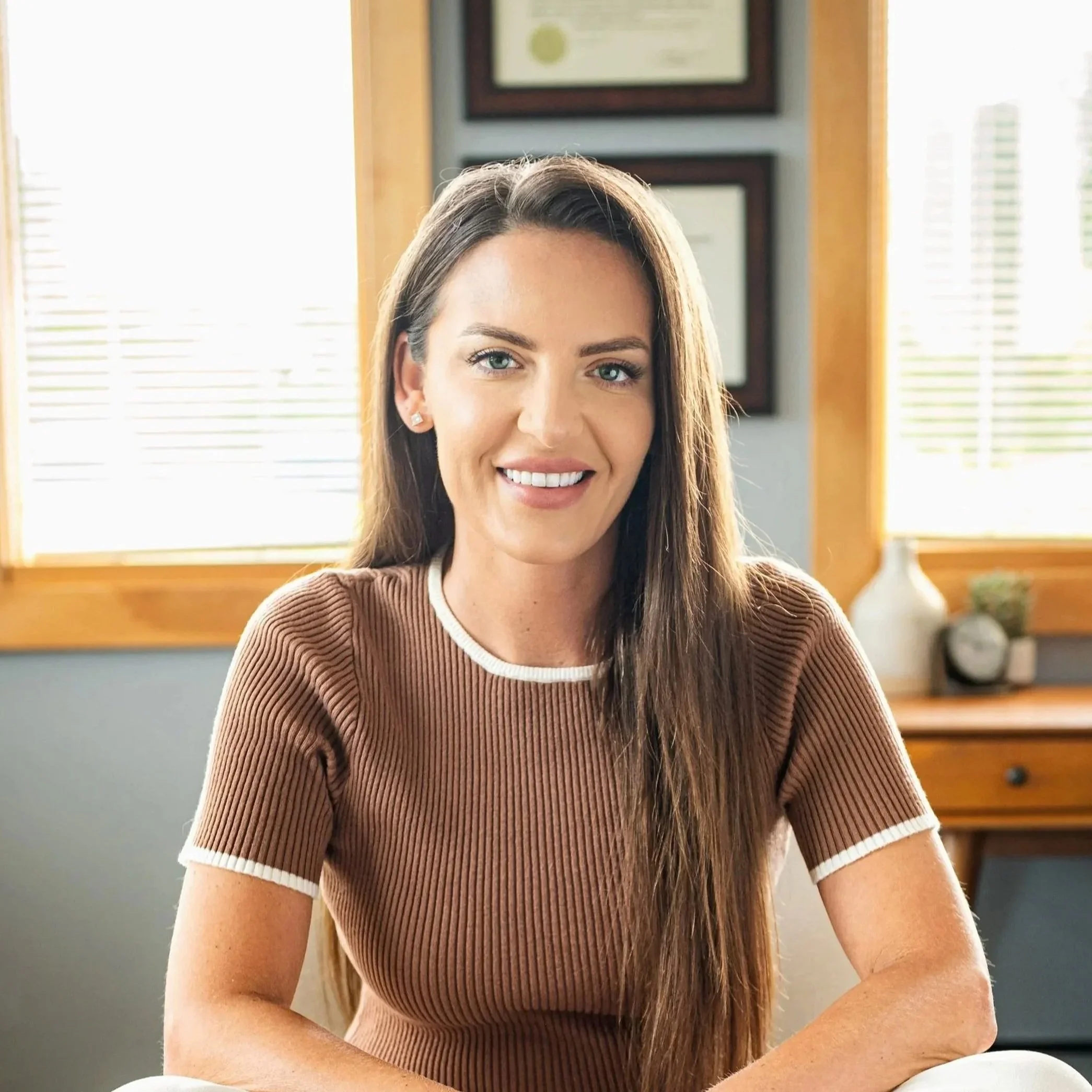 Portrait of a young woman with long brown hair, blue eyes, wearing a beige top with black trim, smiling against a plain background.