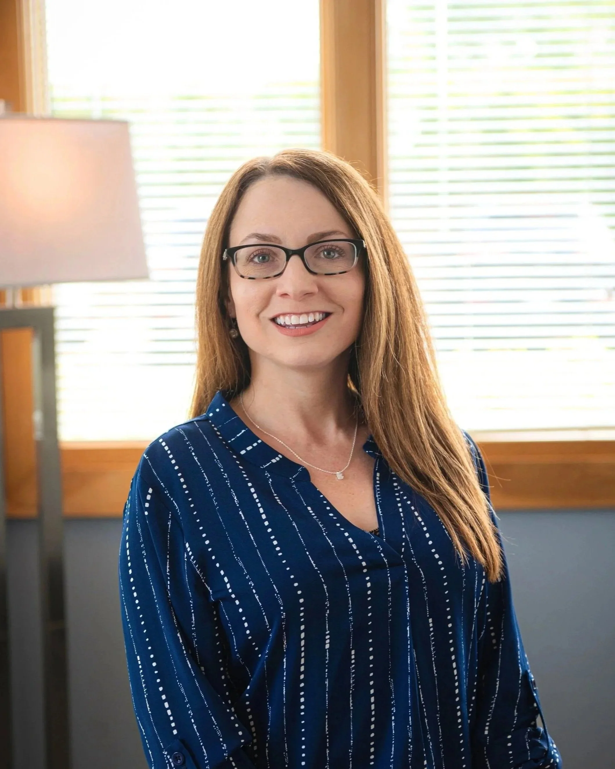 A woman with long brown hair, glasses, and a friendly smile, wearing a gray blouse, standing against a plain beige background.