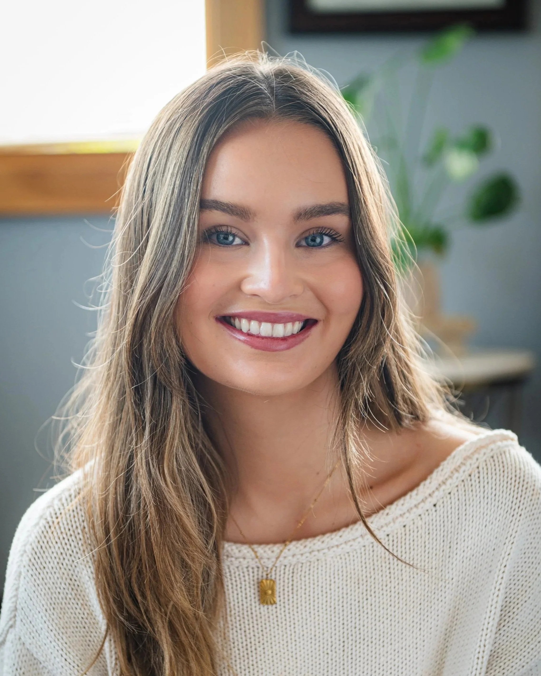 A young woman with long, straight brown hair and blue eyes smiling at the camera, wearing a light green shirt over a white top, against a plain light-colored background.