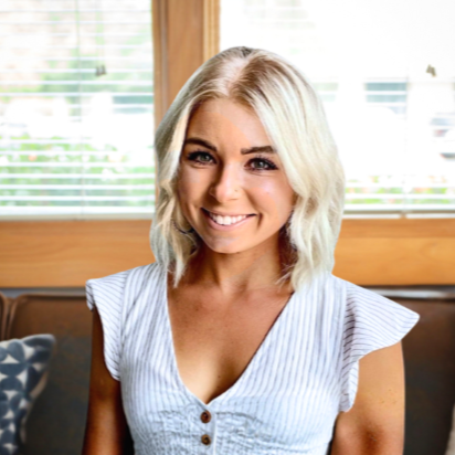 A young woman with blonde, wavy hair smiling at the camera against a plain beige background.