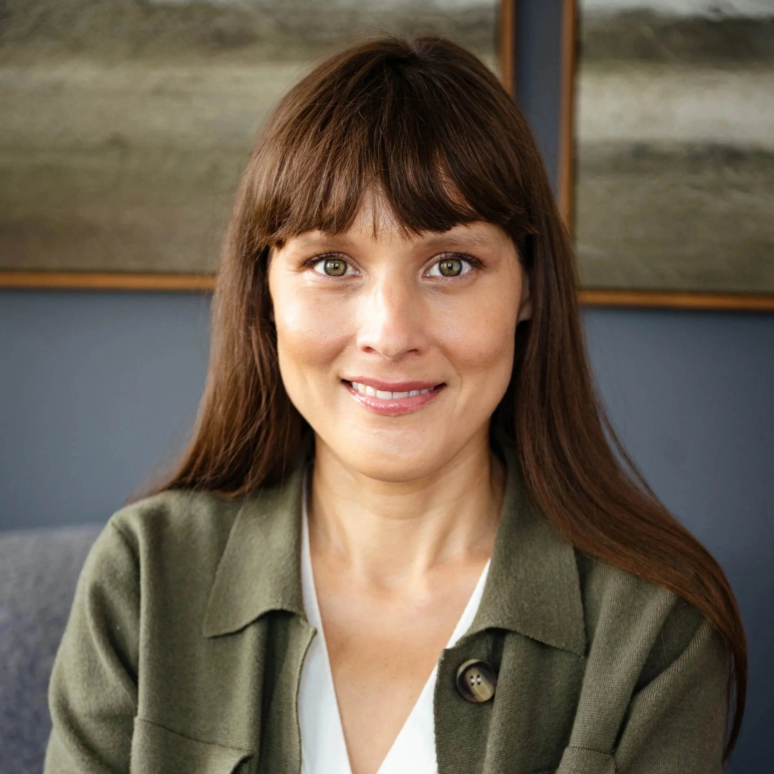 A woman with long brown hair and bangs smiling, wearing a green top, standing against a plain background.