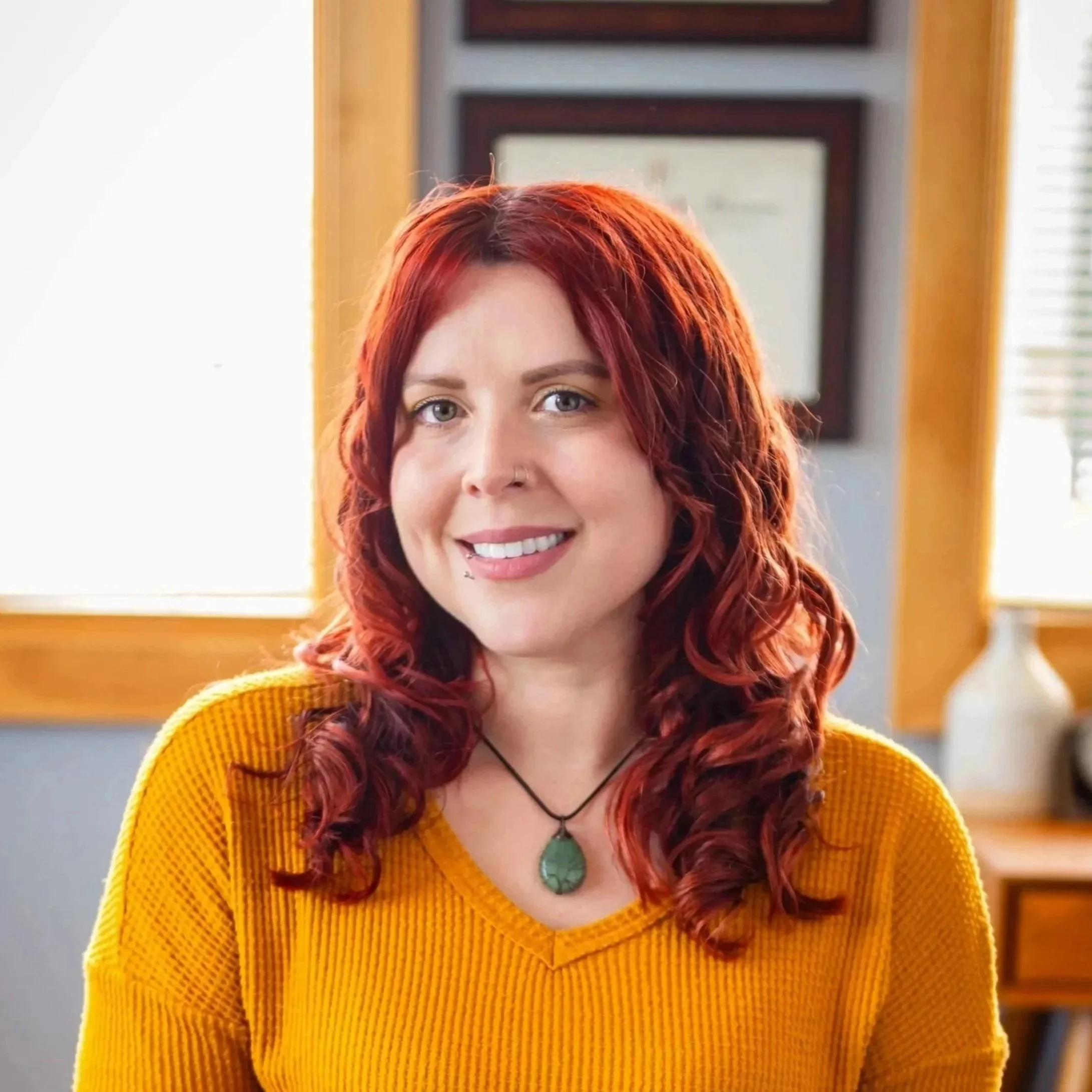 A young woman with curly brown hair, light skin, and green eyes, wearing a nose and lip piercing, a black choker with a green pendant, and an orange sweater, smiling at the camera against a plain background.