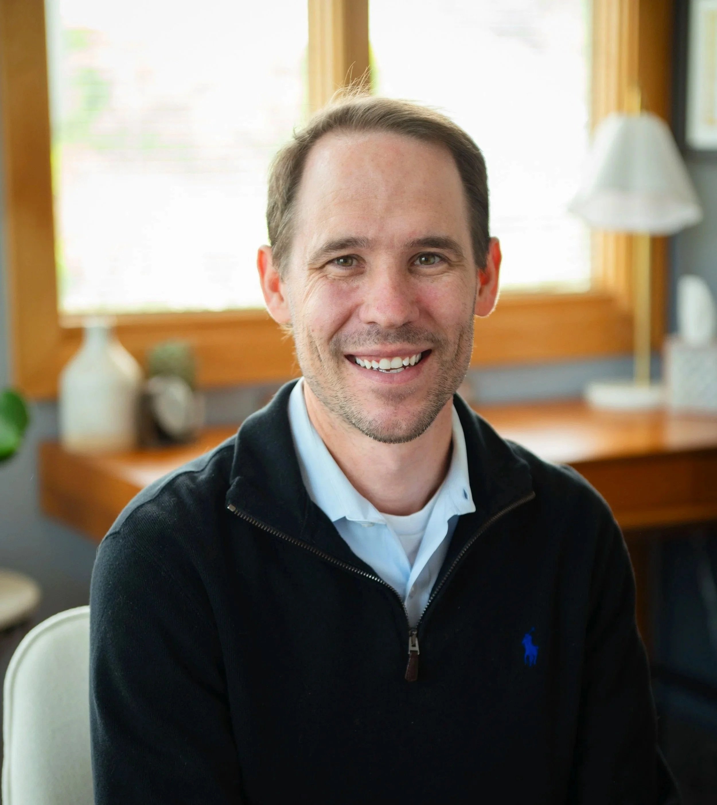 A smiling man with dark hair wearing a checkered shirt and a dark zip-up sweater.
