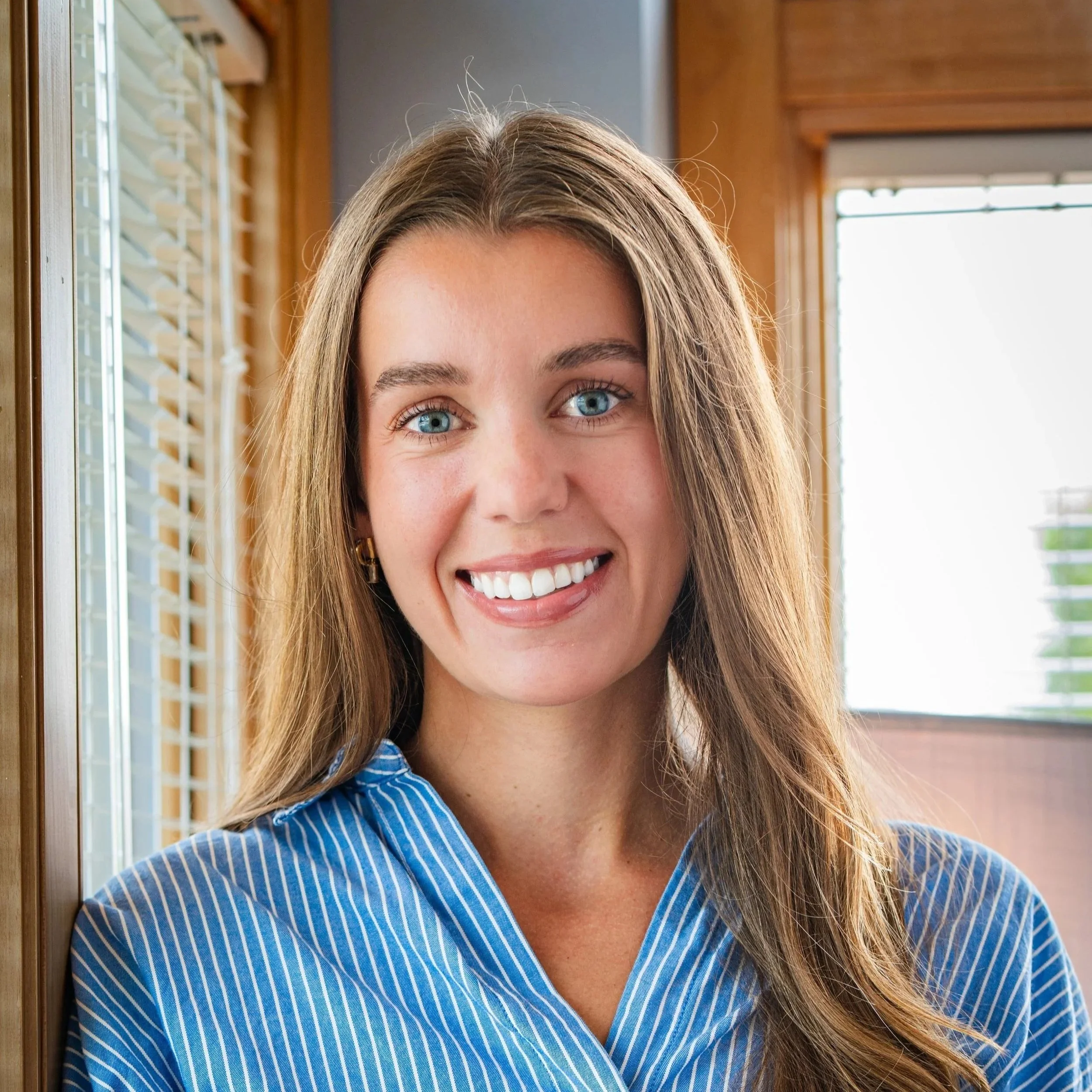 A young woman with long, wavy light brown hair, blue eyes, and fair skin, smiling at the camera, wearing a black top and a checkered blazer, standing against a plain beige background.