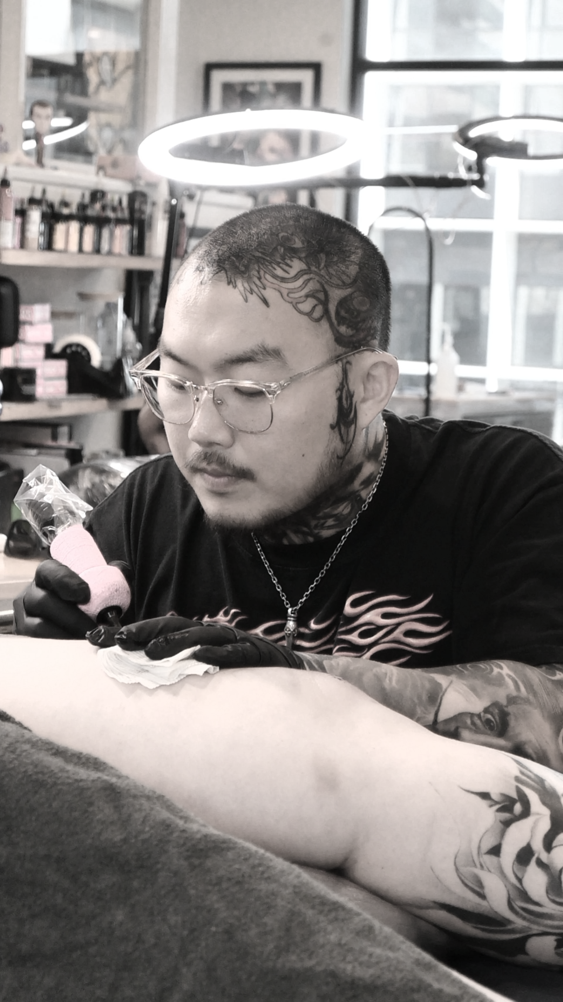 Tattoo artist wearing glasses and gloves tattooing a person's arm in a tattoo studio with shelves and artwork in the background.