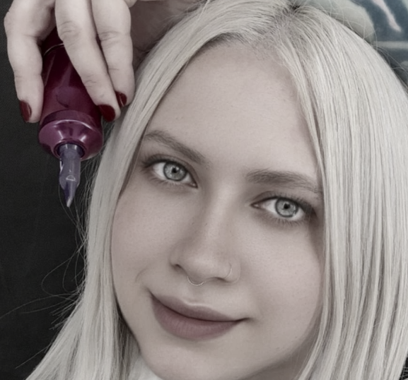 A woman with platinum blonde hair and light blue eyes getting her hair dyed with purple dye, held by a stylist's hand.