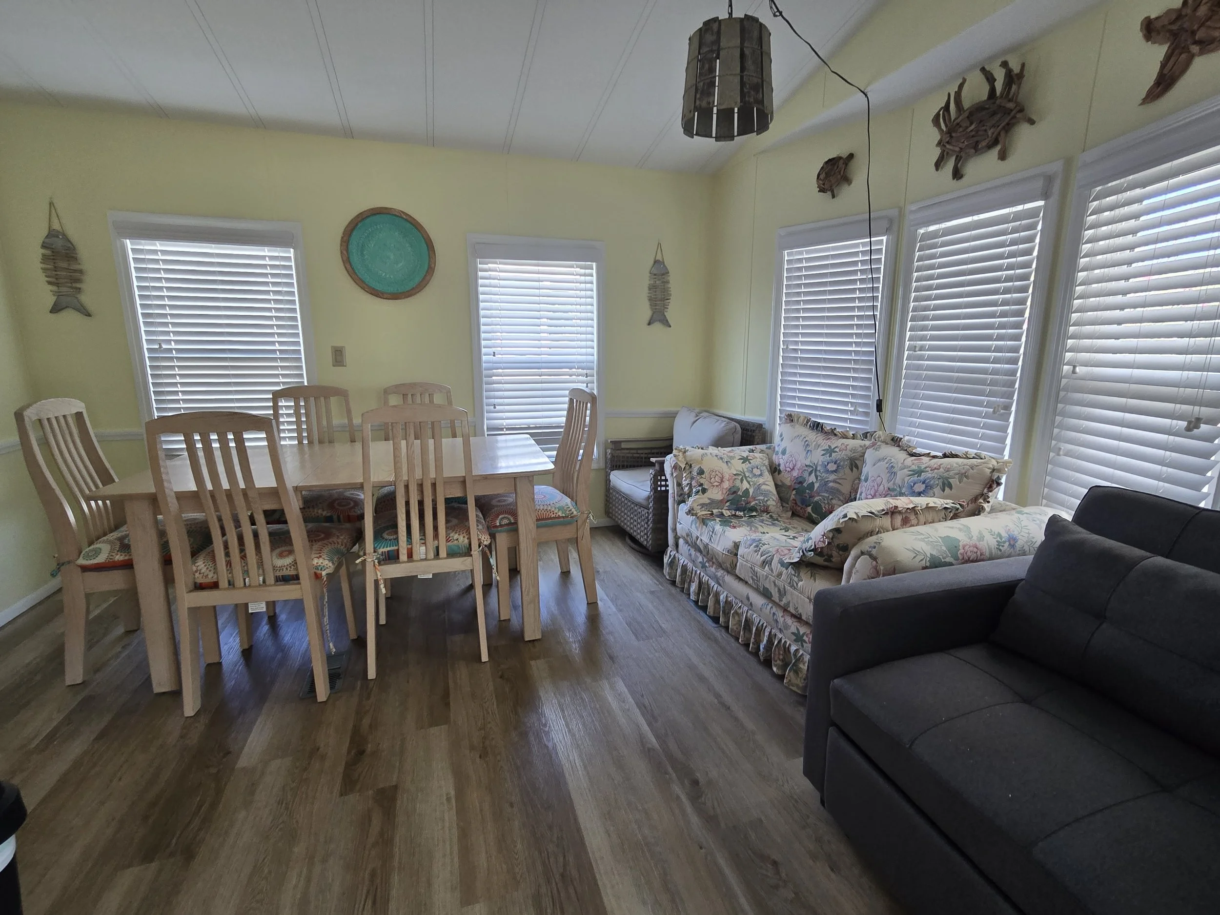 A living room with yellow walls, large windows with white blinds, a round wooden dining table with matching chairs, a floral sofa, a black armchair, and nautical decorations including fish and a turtle on the walls.