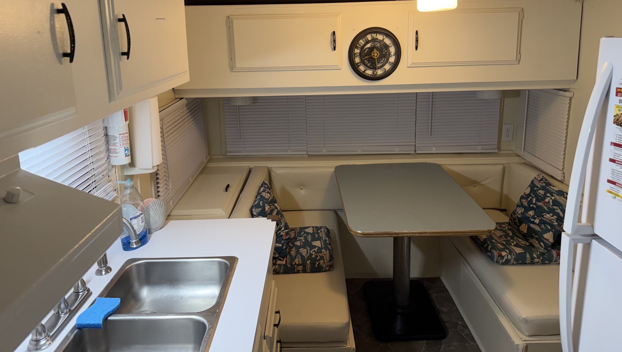 A small kitchen and dining area with beige cabinets, a sink, a chair with patterned upholstery, and a window with closed blinds. There is a clock on the wall, a fire extinguisher, and some items on the countertop.