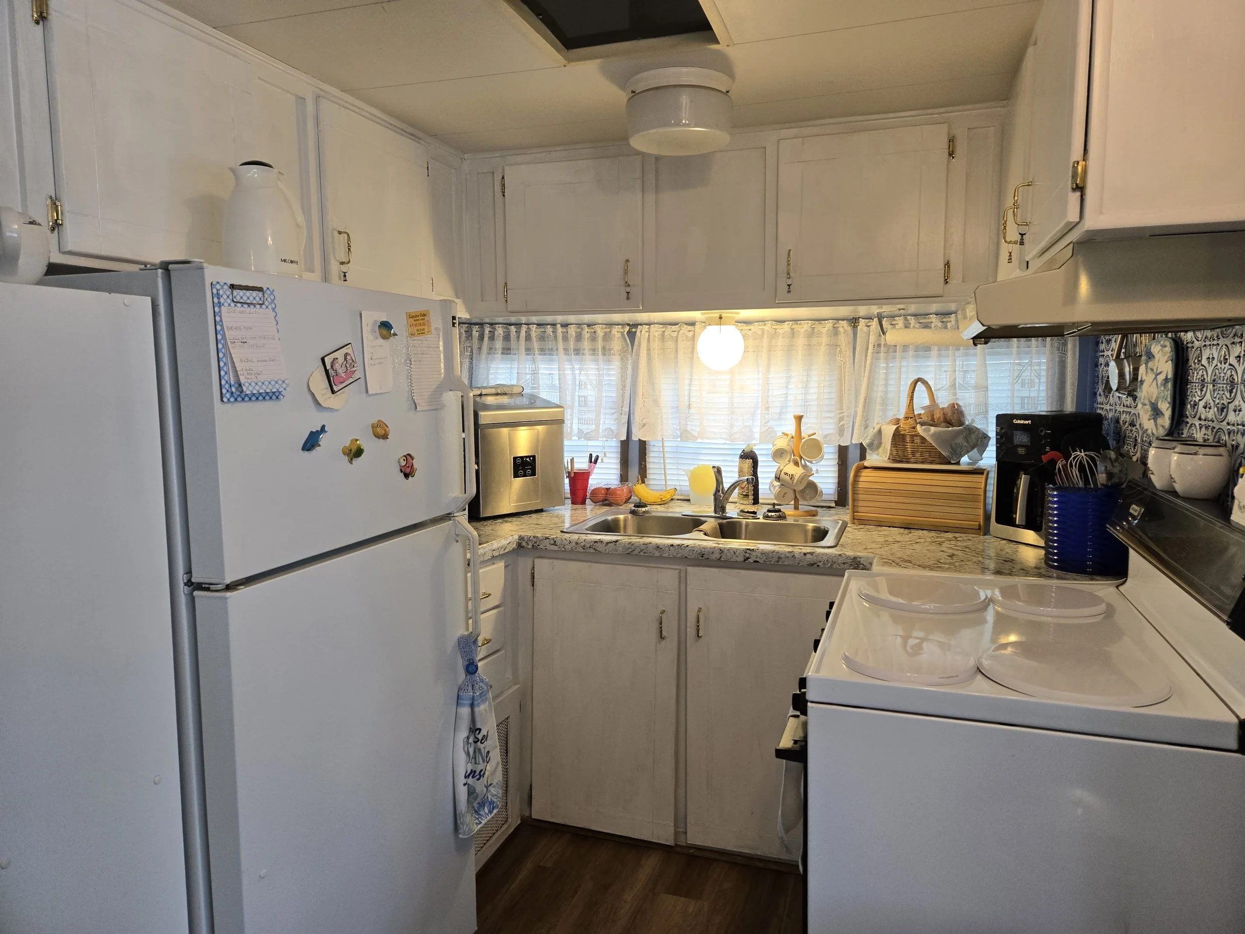 A small kitchen with white cabinets, a white refrigerator with magnets, a granite countertop, a double sink, a coffee machine, and various kitchen items. There is a window with sheer curtains above the sink, and a ceiling light.