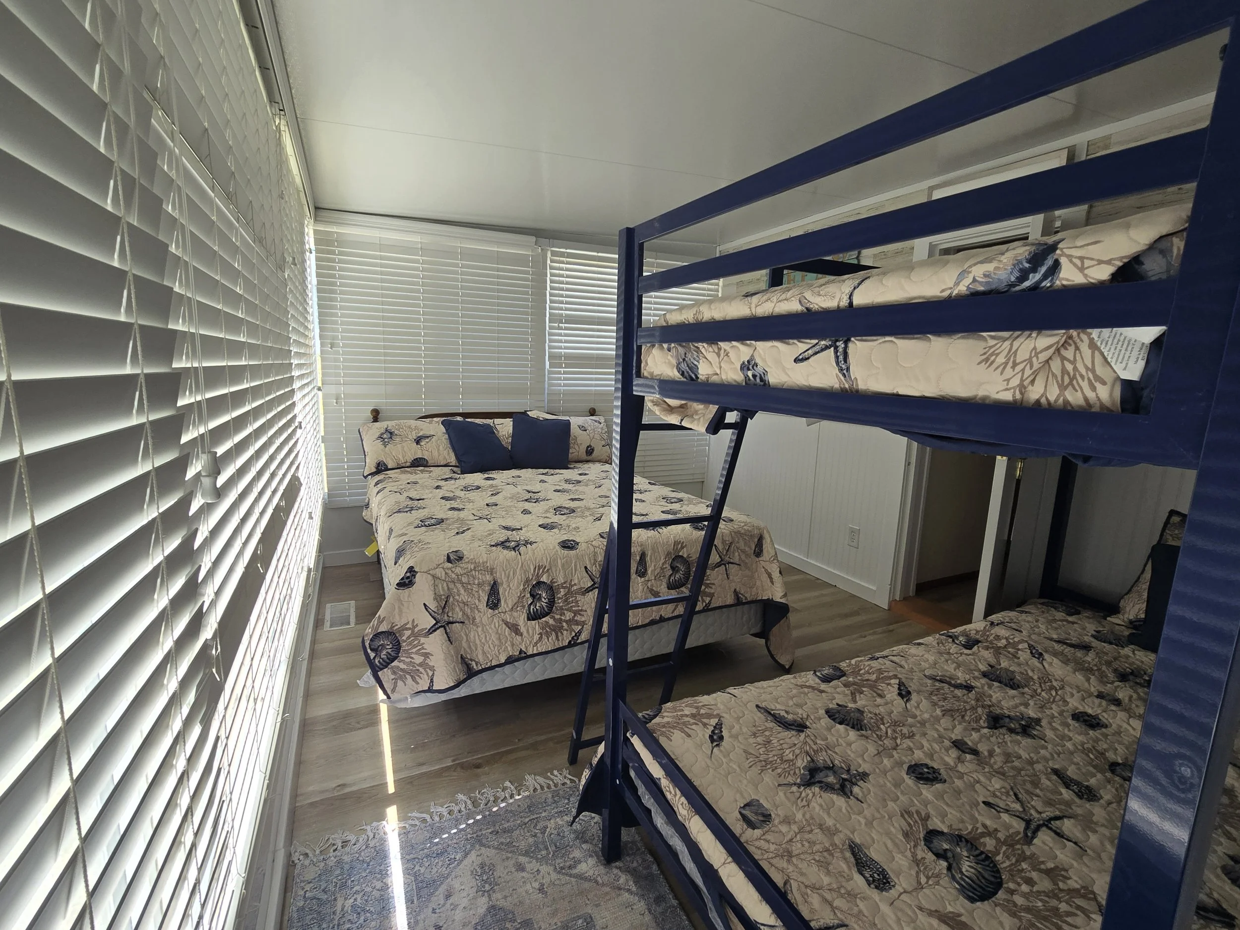 A room with white horizontal blinds on large windows, beige bedding with navy shell and coral reef patterns on a mattress, blue bunk bed frame, carpet with a floral pattern, and a small armchair with matching bedding.