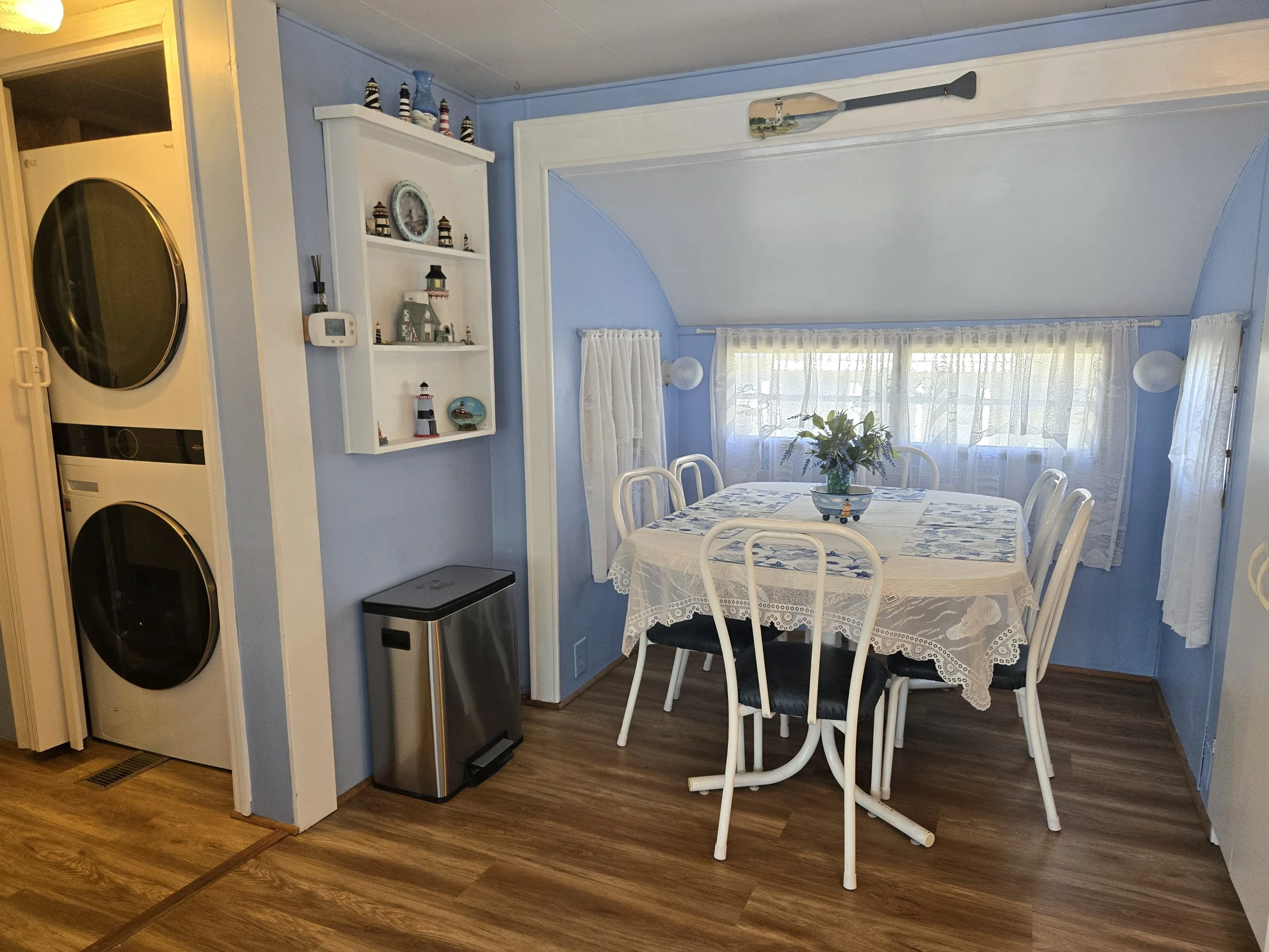 A dining area with a round table covered with a lace tablecloth, six white chairs with black seats, and a vase of flowers on the table. The room has blue walls, a window with lace curtains, and a decorative oar hanging above the window. To the left, 