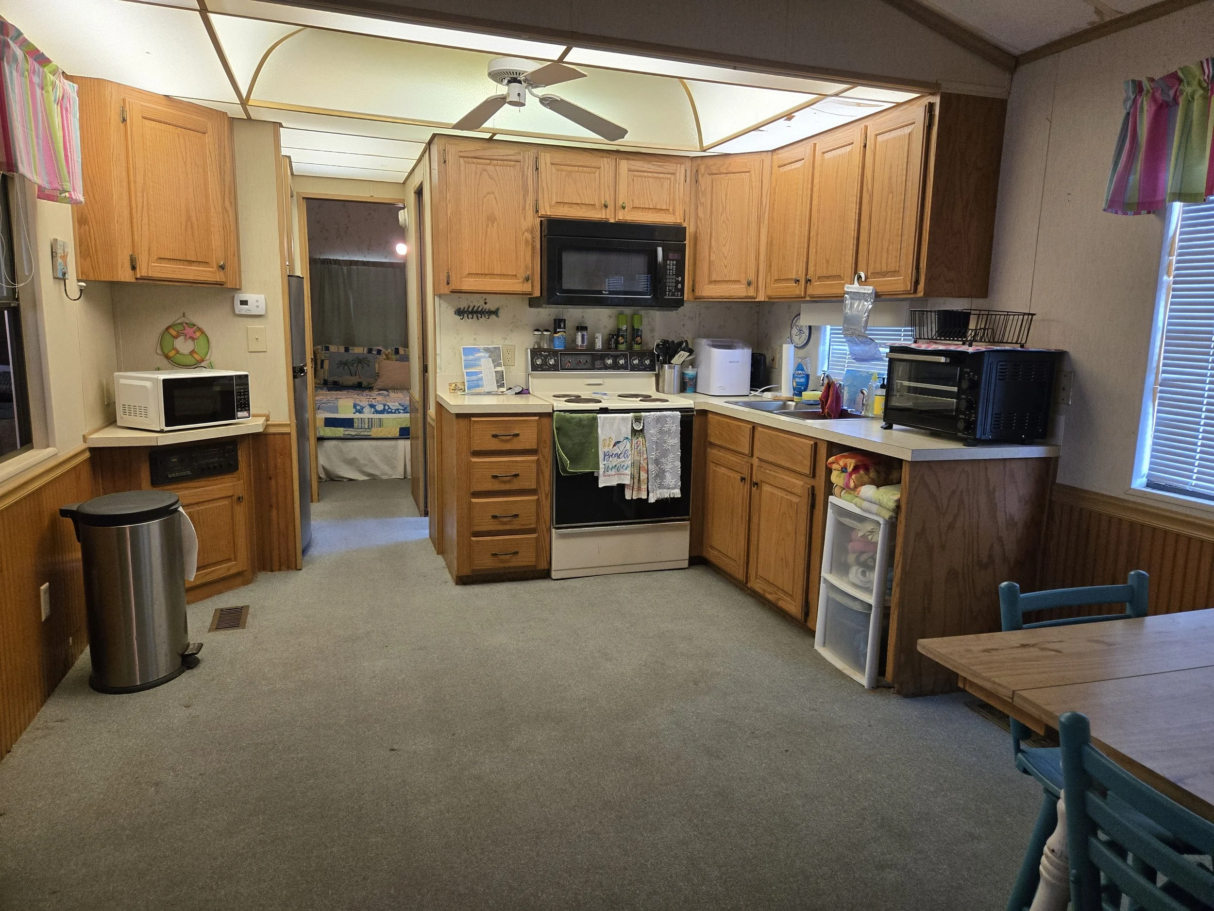 Kitchen with wooden cabinets, white countertop, black microwave, oven, and toaster oven, white sink, window with blinds, small plastic drawer unit, table with teal chair, ceiling fan, beige walls, carpeted floor, and doorway leading to a bedroom.